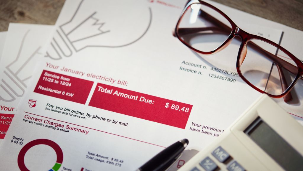 A close-up of an electric bill on a table with a pen, a calculator, and a pair of reading glasses close by.