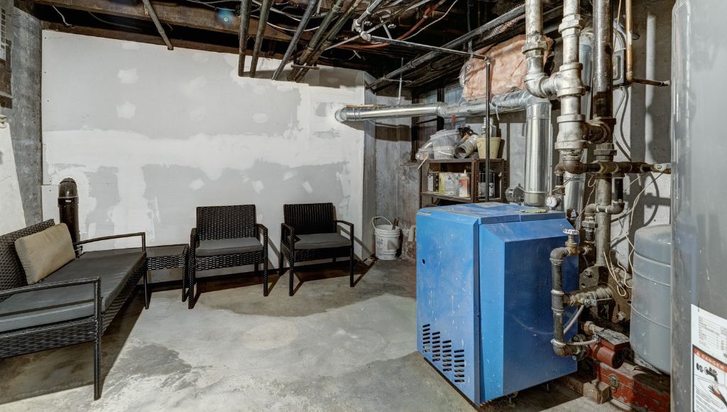 An older furnace and hot water heater in an unfinished basement. Pipes cover the walls, and wicker furniture sits in the corner.