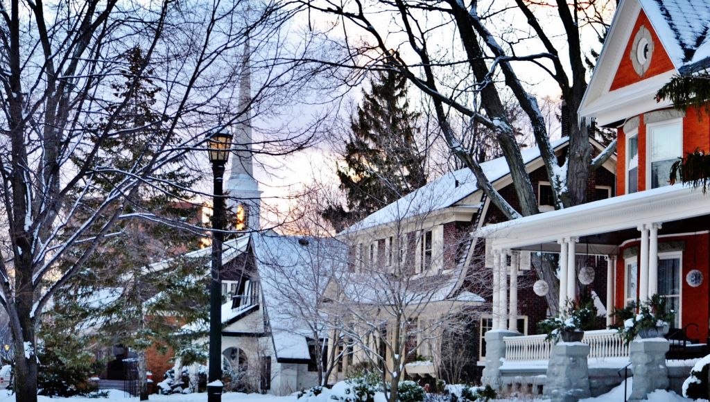 A snowy suburban street in winter shows three houses: a three-story red house, a two-story brown brick house, and a one-story white modern house.