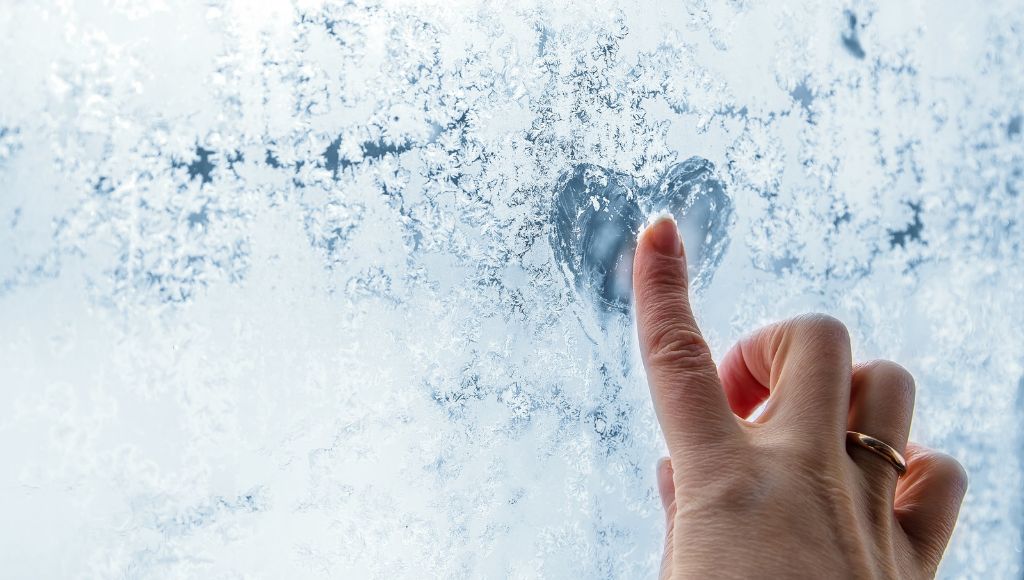 A finger drawing a heart in the frost on the interior of a window. The frost indicates that cold air is getting into the house somewhere.
