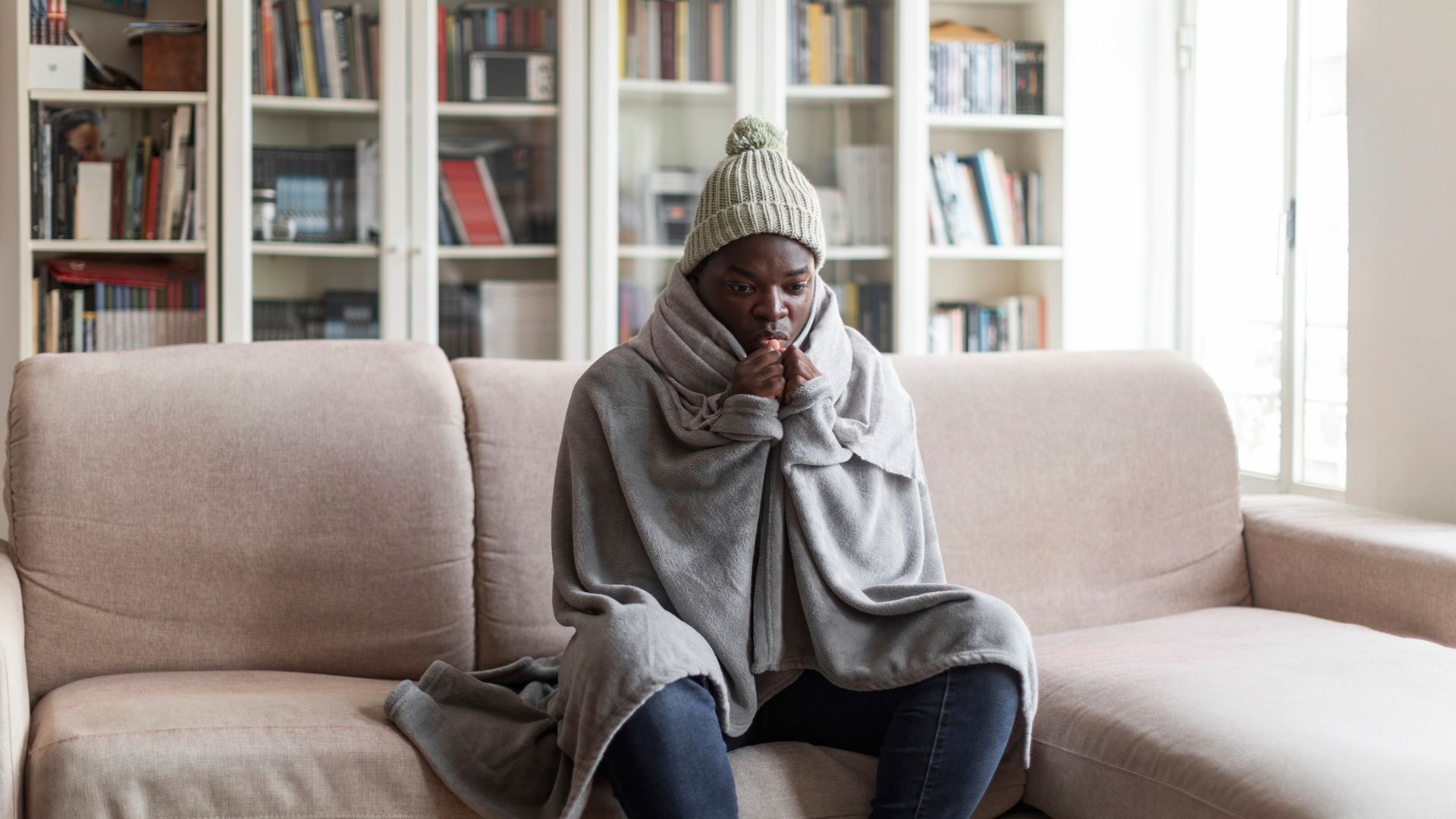 roof-restoration A man sits on his couch in a cold house, wrapped in a blanket and wearing a beige pompom-topped beanie.
